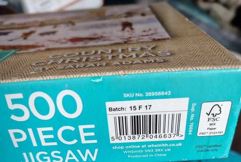 A close-up of a jigsaw puzzle box showing the front and side panels. The box is turquoise and brown, with large white text reading '500 PIECE JIGSAW' and 'COUNTRY CHRISTMAS BY SARAH ADAMS' on the top.