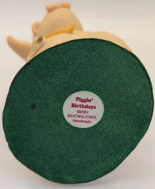 A close-up of the bottom base of a 'Piggin' Birthdays' handmade collectible figure, showing a dark green circular textured base with a white circular sticker in the center.
