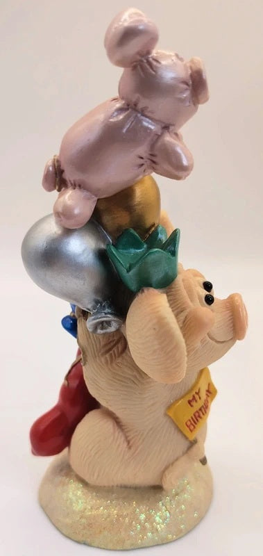 A decorative figurine of a pig stacking balloons, with a pink pig on top, a silver balloon, a green crown, a red boot, and a small yellow sign, set against a plain light background.