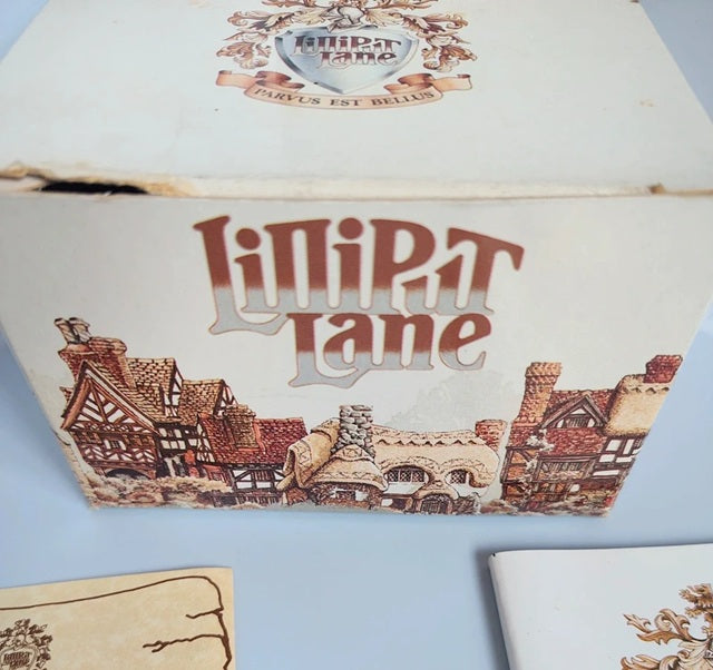 A vintagebox for 'Liliput Lane' featuring an illustration of medieval houses and a crest on the lid, photographed on a light blue surface.