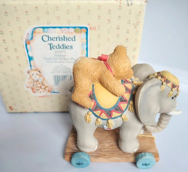 A Cherished Teddy's 'Elephant' figurine carrying a teddy bear on its back, shown with its original packaging box. The figurine features a light gray elephant on a wooden base with blue wheels, decorated with a patterned saddle and a golden-brown teddy bear carrying a pink bow.