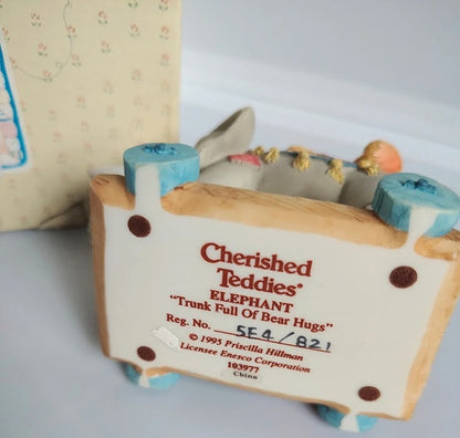 A collectible ceramic figurine of an elephant in a trunk, labeled 'Cherished Teddy's ELEPHANT "Trunk Full Of Bear Hugs"', with a patterned box in the background.