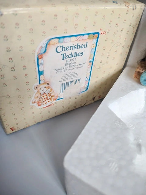 A package of Cherished Teddies plush toys, showing the cream-colored box with a teddy bear illustration and label, next to a white plastic bag.