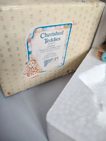 A package of Cherished Teddies plush toys, showing the cream-colored box with a teddy bear illustration and label, next to a white plastic bag.