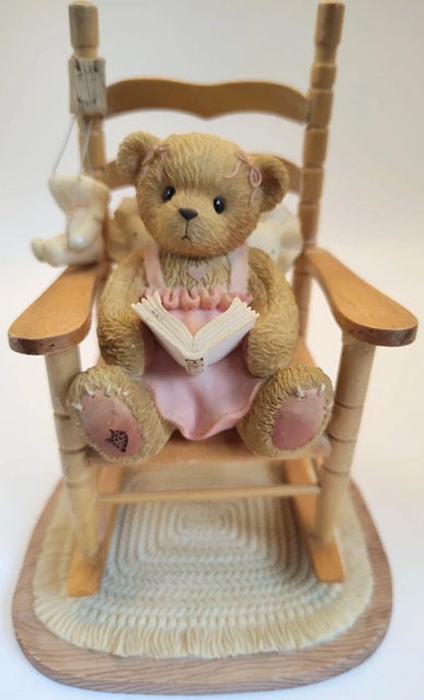 Cherished Teddies Lucy teddy bear in a pink outfit sitting on a wooden rocking chair with a white background