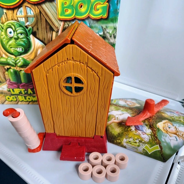 Toy house with cylindrical objects on a tray, with a game box featuring cartoon characters in the background.