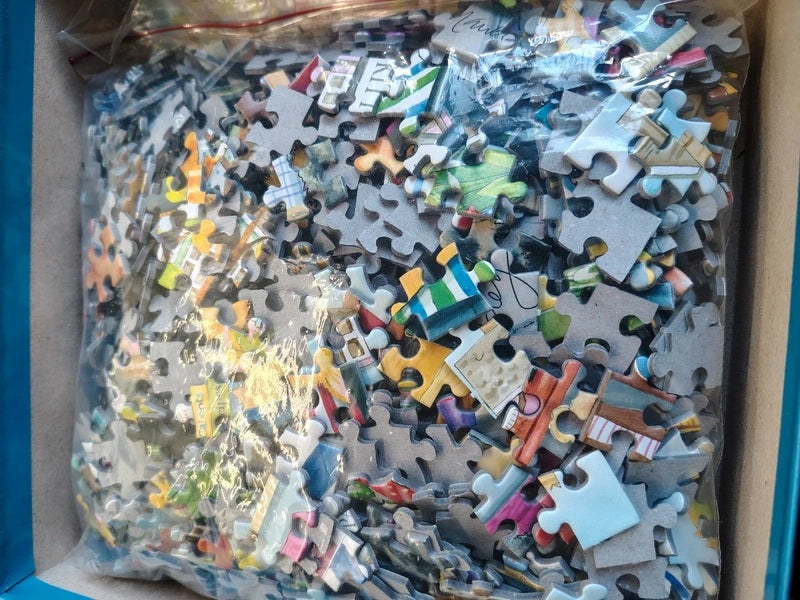 A box of loose jigsaw puzzle pieces in a clear plastic bag, showing many small puzzle pieces in various colors and shapes.