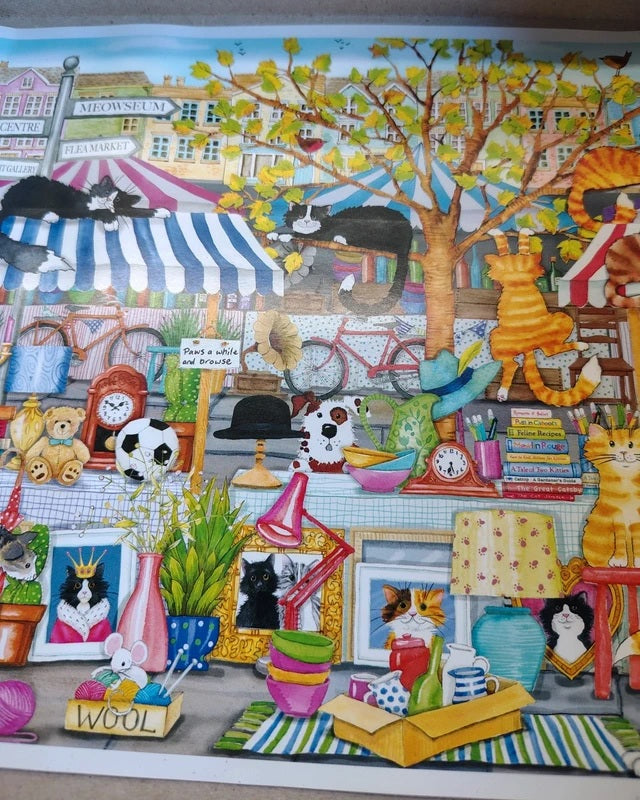 A colorful illustrated scene showing a busy market or shop display with many cats, framed art, books, baskets, plants, and striped awnings. The artwork appears to be the product itself rather than a photo of a physical object in a room, so this fits Scenery.