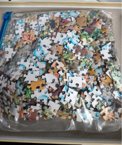 A clear plastic zippered pouch filled with assorted jigsaw puzzle pieces, photographed from above inside a tray or box.
