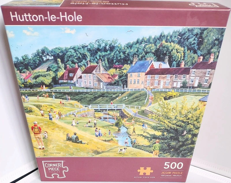 A 500-piece jigsaw puzzle box for Hutton-le-Hole, featuring a colorful illustration of a village scene with houses, trees, and people by a stream. The box has a dark red border with the title 'Hutton-le-Hole' at the top and the 'Corner Piece' brand logo at the bottom left.