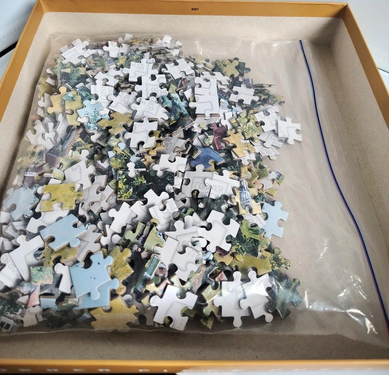 A box of loose jigsaw puzzle pieces sealed in a clear plastic bag, photographed from above inside a tan cardboard box.