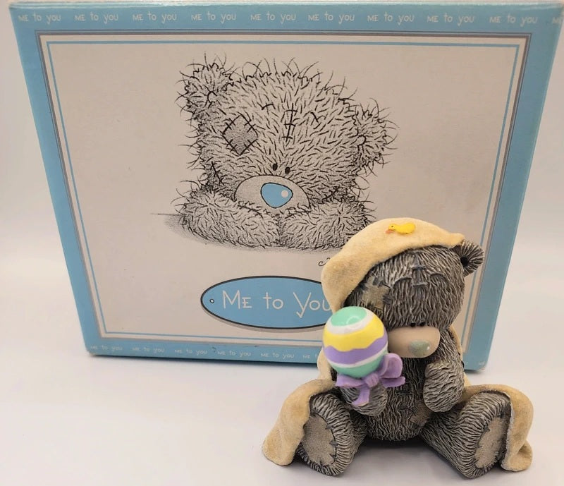 A Me to You  Baby Love teddy bear figurine sitting in front of its original light blue and white box. The bear is gray with a beige hat and is holding a small pastel-colored striped rattle.