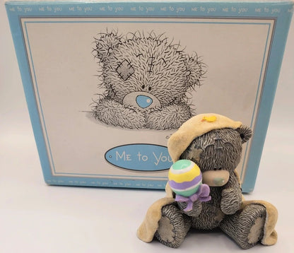 A Me to You  Baby Love teddy bear figurine sitting in front of its original light blue and white box. The bear is gray with a beige hat and is holding a small pastel-colored striped rattle.