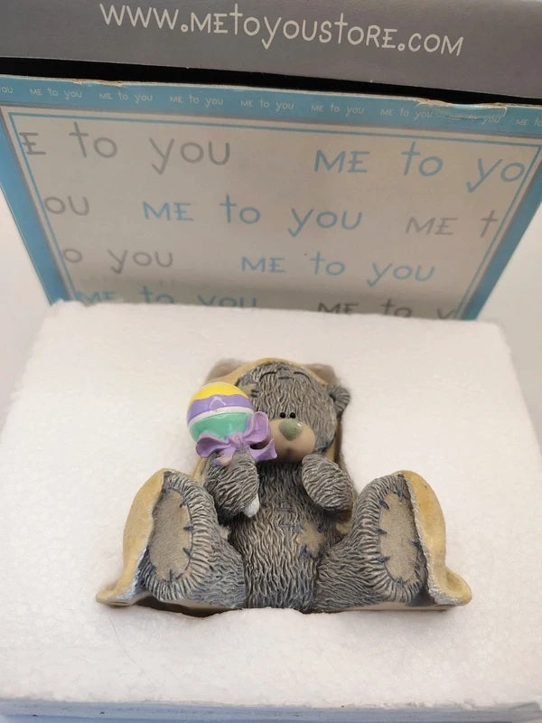 A small teddy bear figurine sitting in a white foam insert, holding a pastel rainbow lollipop, with its original blue and white packaging box behind it.