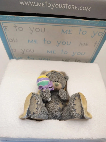 A small teddy bear figurine sitting in a white foam insert, holding a pastel rainbow lollipop, with its original blue and white packaging box behind it.