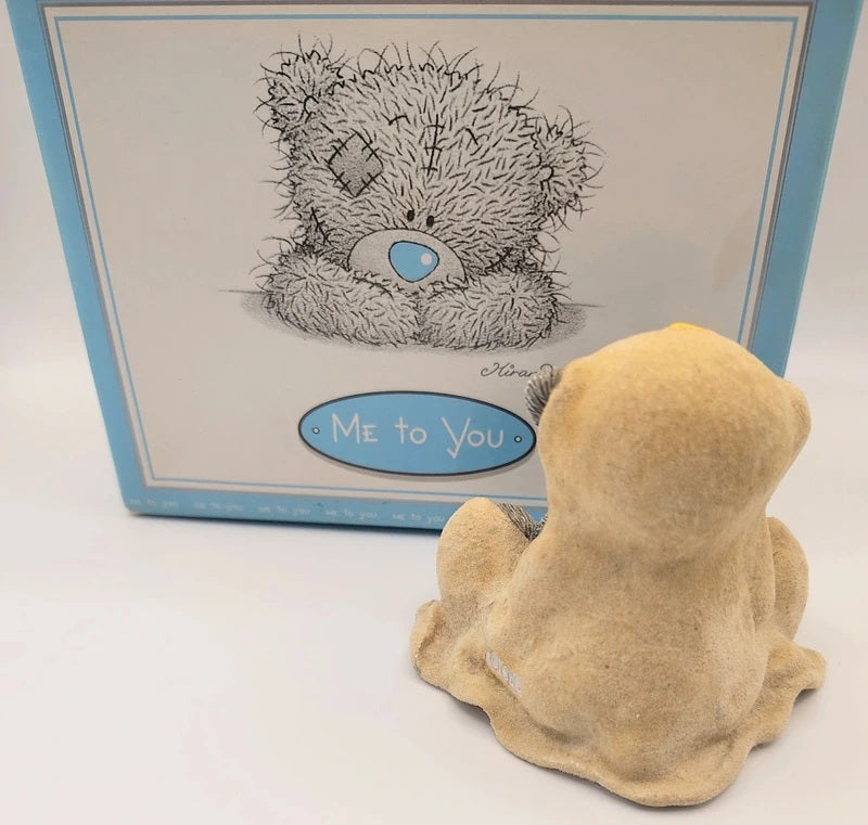 A Me to You teddy bear figurine shown from the back in a light tan color, positioned in front of its original blue and white packaging box featuring a black-and-white illustration of the bear and the text 'Me to You'.