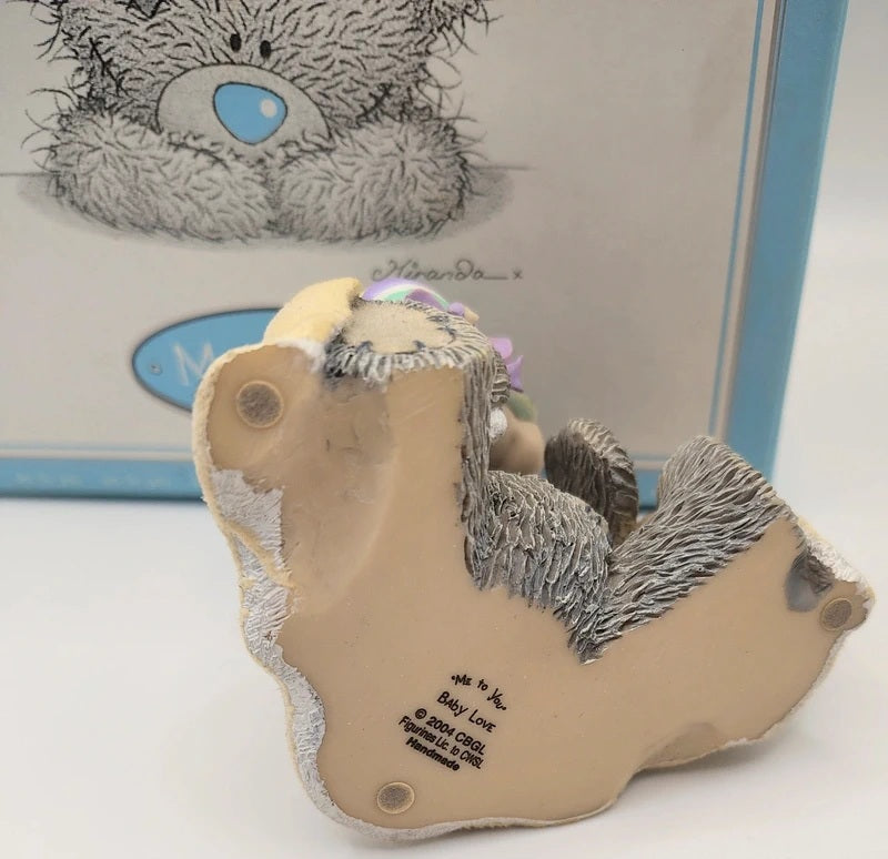 A Me Too Bear collectible figurine shown from its underside, featuring a beige body with gray and black textured fur details, set against a blurred background of its original packaging.