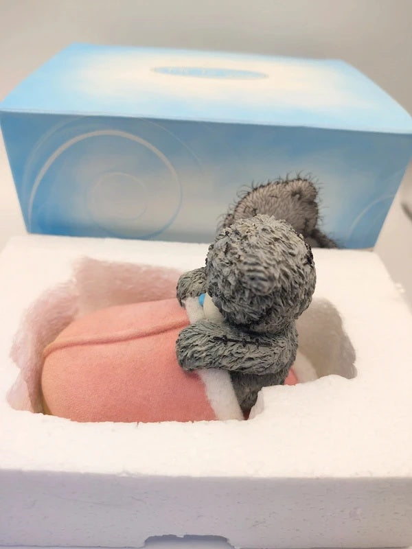 A small gray resin figurine hugging a pink oval object inside a white foam insert, with a blue-and-white product box behind it.