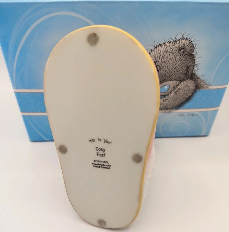 A Me To You 'Cosy Feet' collectible ceramic figurine shown from its back, displaying the white glazed base with printed text and four grey dots, with its blue illustrated packaging box in the background.