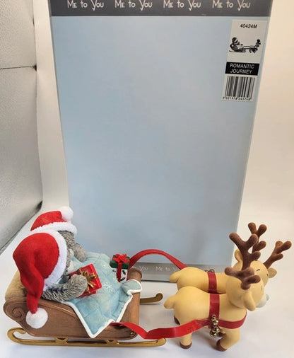 A Christmas-themed ornament or figurine featuring a red Santa Claus sitting in a sleigh pulled by two tan reindeer with brown antlers, set in front of a large light blue package card.