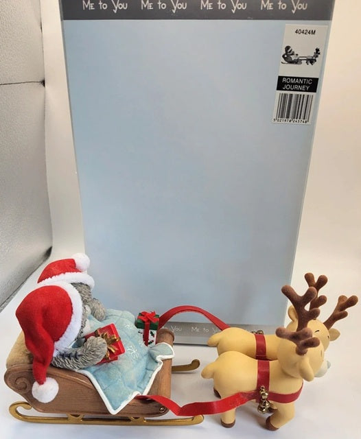A Christmas-themed ornament or figurine featuring a red Santa Claus sitting in a sleigh pulled by two tan reindeer with brown antlers, set in front of a large light blue package card.
