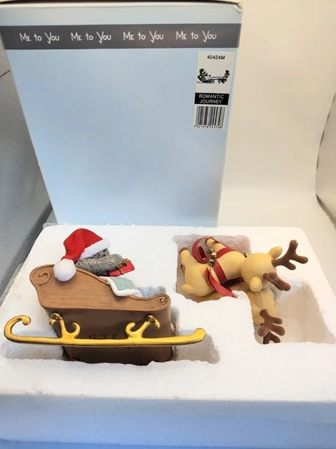 A Christmas ornament set in its original packaging, featuring a brown sleigh with gold trim and a red Santa hat, alongside a brown reindeer with a red and gold harness. The set is displayed in white foam insert in front of its light blue and white box.