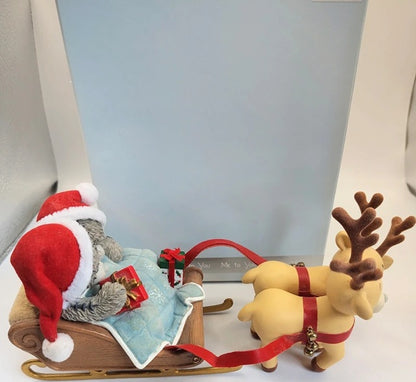 A Christmas-themed ornament featuring a gray cat wearing a red Santa Claus and sitting in a wooden sleigh pulled by two tan reindeer with red harnesses, set against a light blue box.