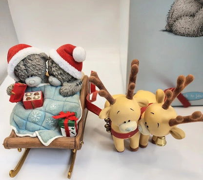 A Christmas ornament or figurine set featuring two gray teddy bears wearing Santa hats in a wooden sleigh with gifts, pulled by two yellow reindeer with brown antlers and red harnesses, displayed on a white surface.