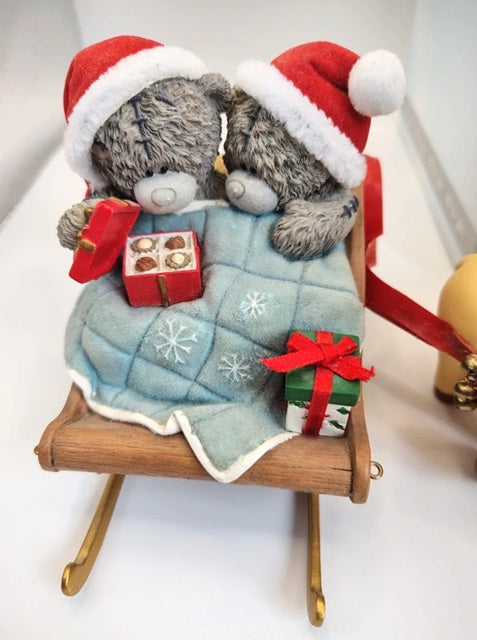 A holiday ornament featuring two grey teddy bears wearing red Santa hats, sitting in a wooden sleigh with a light blue quilted blanket decorated with white snowflakes. One bear holds a small red gift box, and the sleigh is shown on a white surface with a small green gift box in the foreground.