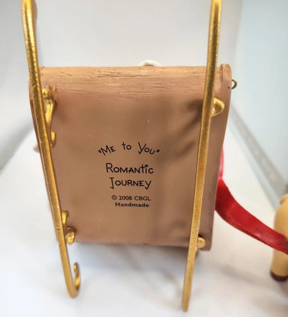 A handmade ornament shaped like a sleigh with gold metal frame-like sides, a red ribbon handle, and handwritten text reading “Me to You Romantic Journey” with “© 2008 CBGL Handmade” on the cover, shown on a white surface.