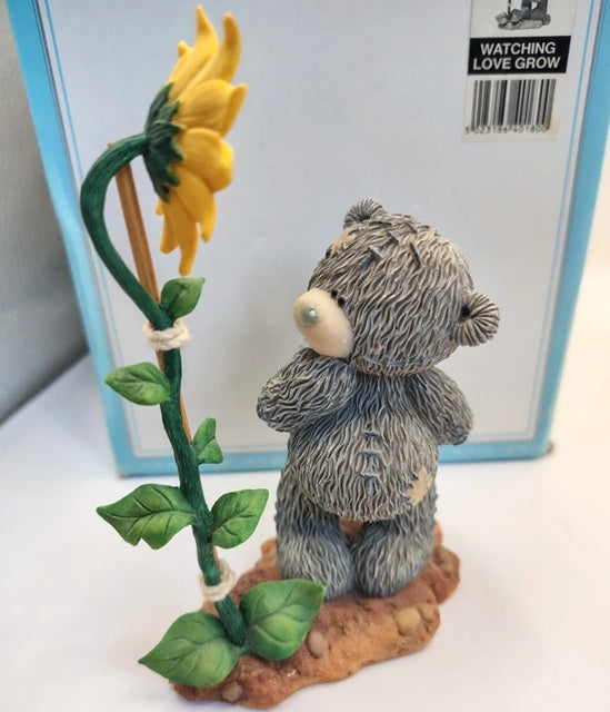 A small gray teddy bear figurine standing on a brown base next to a yellow sunflower with green leaves, in front of a light blue box labeled 'WATCHING LOVE GROW'.