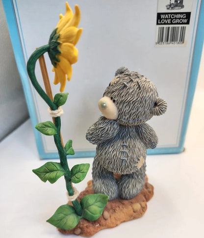 A small gray teddy bear figurine standing on a brown base next to a yellow sunflower with green leaves, in front of a light blue box labeled 'WATCHING LOVE GROW'.