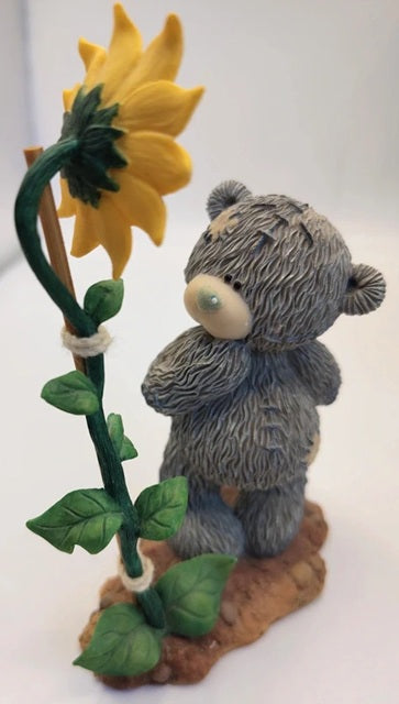 A small gray teddy bear figurine standing beside a large yellow sunflower on a brown base, photographed against a plain light background.
