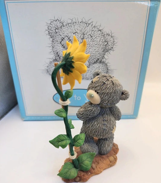 A small gray teddy bear figurine standing beside a yellow sunflower on a brown base, with a light blue box behind it on a white surface.