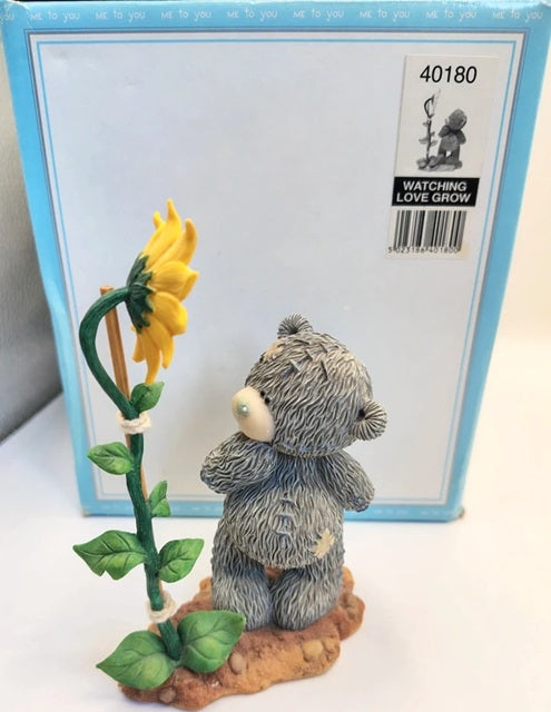 A small gray Me To You  teddy bear figurine standing beside a yellow sunflower on a brown base, placed in front of its light blue retail box. The box shows the text 'WATCHING LOVE GROW' and product number 40180.