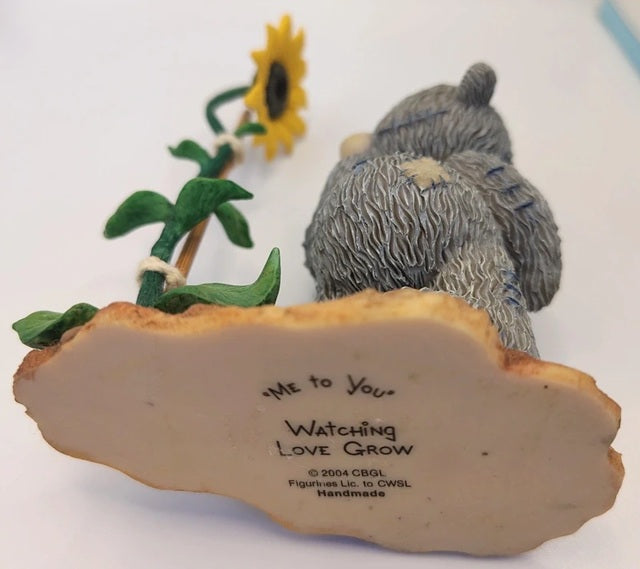 A handmade ceramic figurine of two gray bears sitting on a cloud base with a yellow sunflower, photographed on a white background.