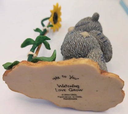 A handmade ceramic figurine of two gray bears sitting on a cloud base with a yellow sunflower, photographed on a white background.