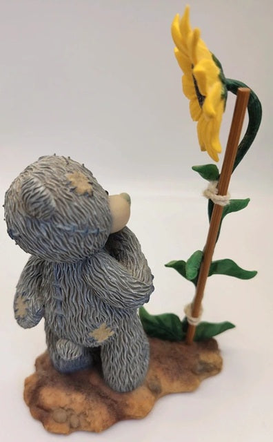 A small gray teddy bear figurine with a stitched look, standing on a brown base next to a yellow sunflower on a white background.