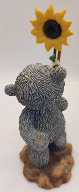 A teddy bear figurine holding a yellow sunflower, standing on a brown textured base against a plain white background.