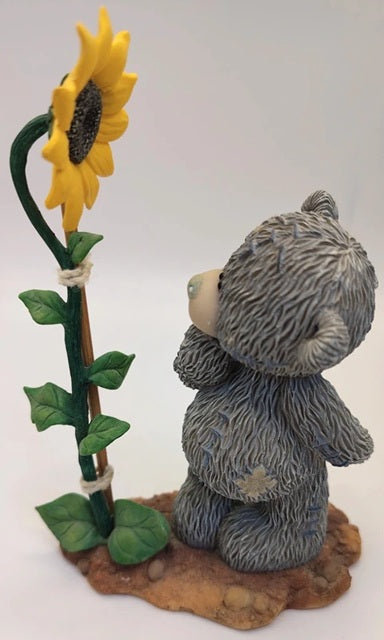 A small gray teddy bear figurine sitting beside a tall yellow sunflower on a brown base against a plain light background.