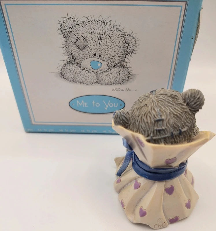 A Me to You teddy bear figurine shown from the back, wearing a cream dress with purple hearts and a blue ribbon, standing in front of its original blue and white box.