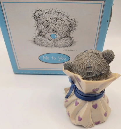 A Me to You teddy bear figurine shown from the back, wearing a cream dress with purple hearts and a blue ribbon, standing in front of its original blue and white box.