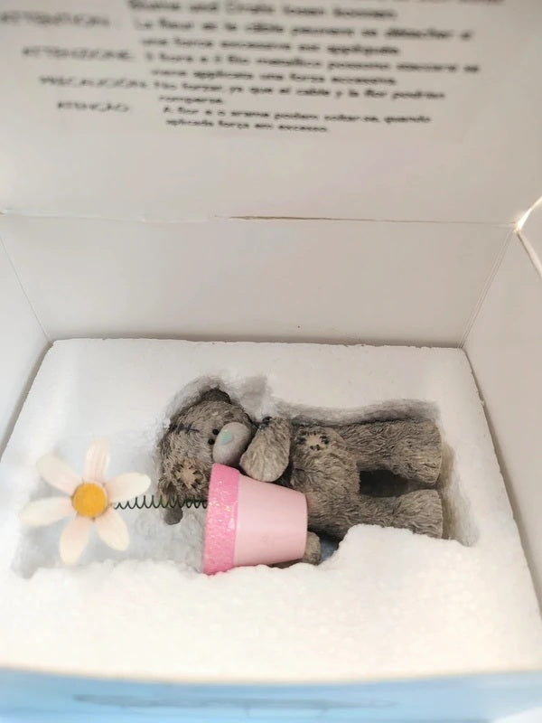A small gray teddy bear figurine wrapped in a pink pot, displayed inside a white gift box with a white foam insert and a daisy flower accent.