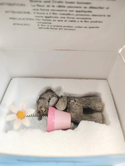 A small gray teddy bear figurine wrapped in a pink pot, displayed inside a white gift box with a white foam insert and a daisy flower accent.