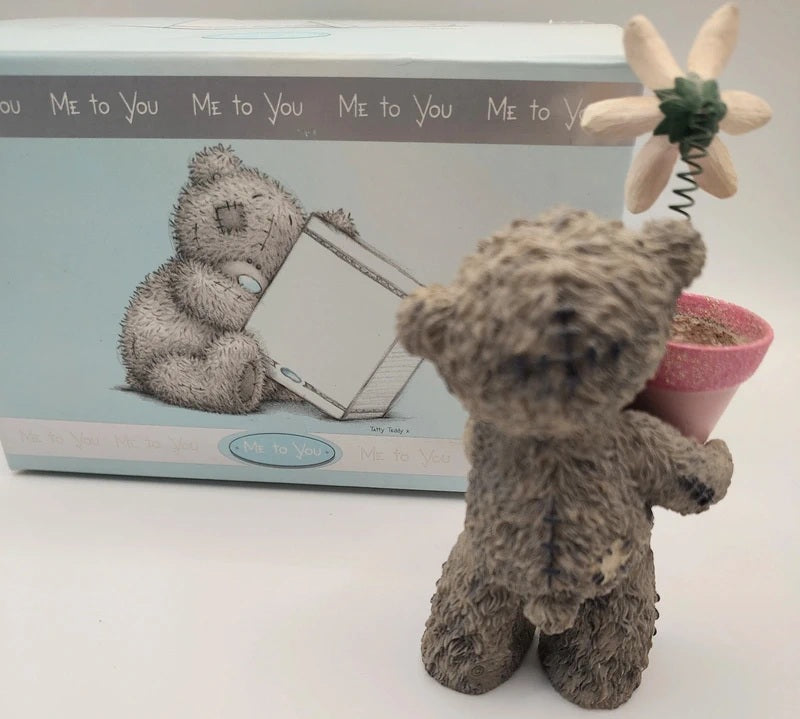 A Me To You teddy bear figurine holding a small pink flower pot with a flower on a spring, shown in front of its light blue branded packaging box.