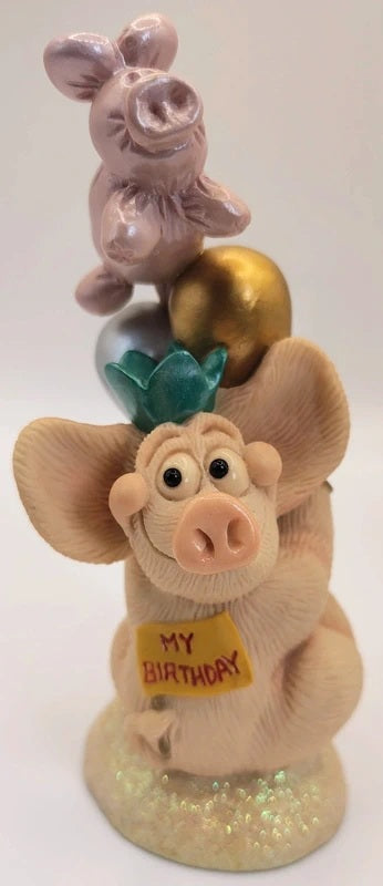 A pig-shaped figurine with a gold ball on its head, a green flower crown, and a yellow 'MY BIRTHDAY' sign, standing on a glittery base against a white background. Piggin Birthdays figurine.