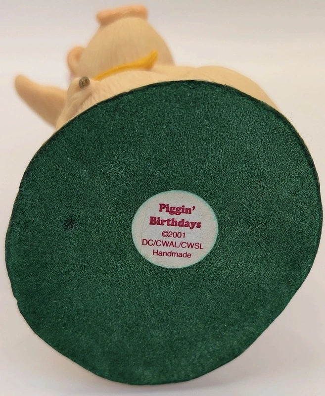 A close-up of the bottom base of a 'Piggin' Birthdays' handmade collectible figure, showing a dark green circular textured base with a white circular sticker in the center.