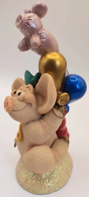 A decorative figurine of a pig character carrying a stack of balloons, including a pink pig on top, a gold balloon, a blue balloon, and a green bow, standing on a glittery base.