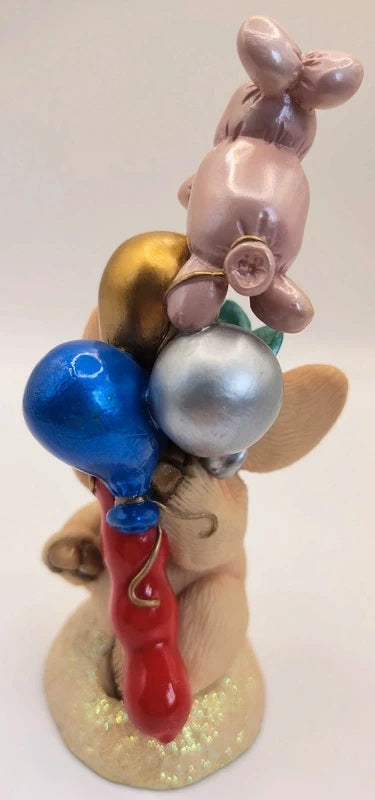 A decorative figurine of a seated figure holding a stack of glossy balloons in blue, silver, gold, and red, on a glittery beige base against a plain light background.