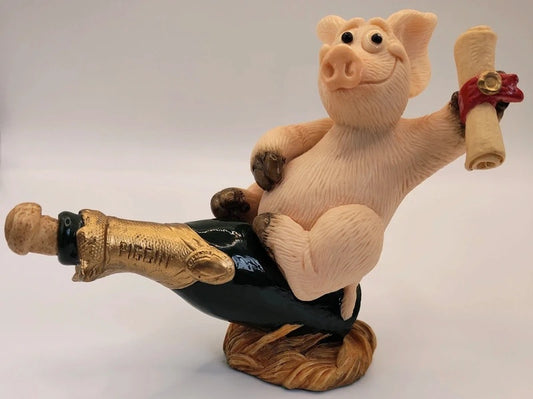 A decorative figurine of a pink pig sitting on a dark bottle with gold foil, holding a rolled scroll with a red ribbon, set on a brown base against a light gray background. Piggin Celebrations figurine.